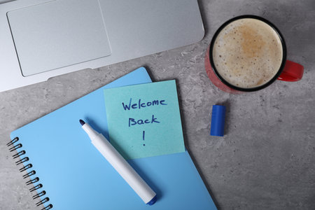 Sticky note with phrase Welcome Back, laptop, coffee, marker and notebook on grey table, flat layの写真素材