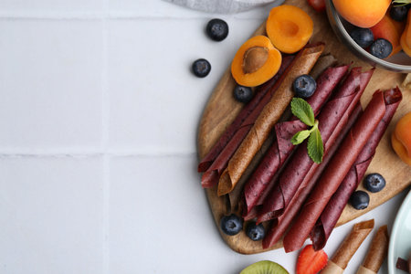 Tasty candied leather rolls, fruits and berries on white tiled table, flat lay. Space for textの写真素材