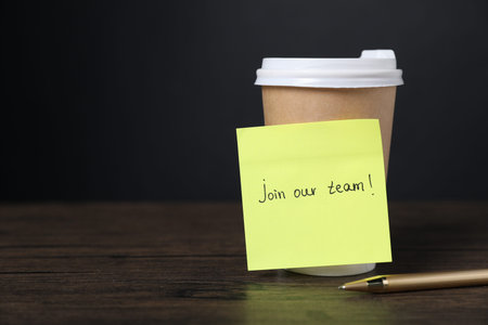 Paper note with the phrase Join Our Team, pen and cup of coffee on wooden table. Space for text