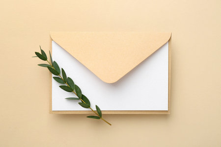 Blank invitation card, envelope and green twig on beige background, top view