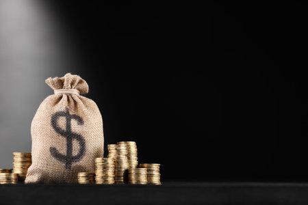 Bag with dollar sign and stacked coins on black table against dark gradient background, space for...