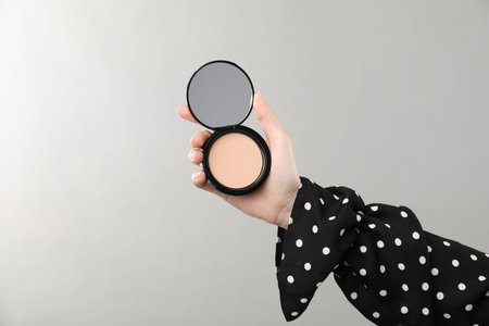 Woman holding open face powder with mirror on gray background, closeup. Space for text