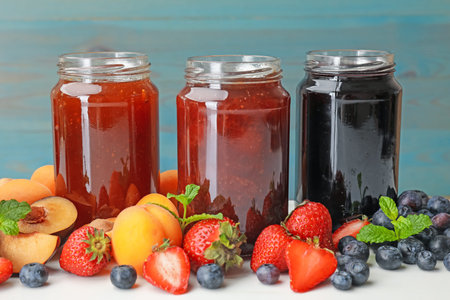 Different jams in jars and ingredients on white table, closeup