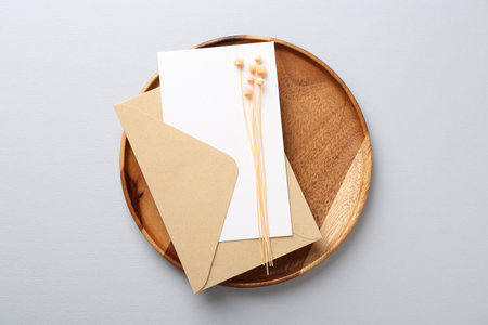 Blank invitation card, envelope, dry spikes and wooden tray on light gray background, top view