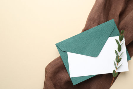 Blank invitation card, envelope and green twig on beige background, top view. Space for text