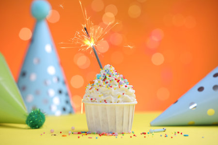 Burning sparkler in cupcake, sprinkles and party decor on yellow table against orange background blurred lightsの写真素材