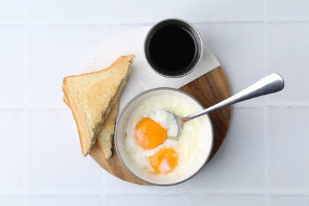 Half-boiled eggs in bowl, soy sauce and toasted bread on white tiled table, flat lay. Traditional...