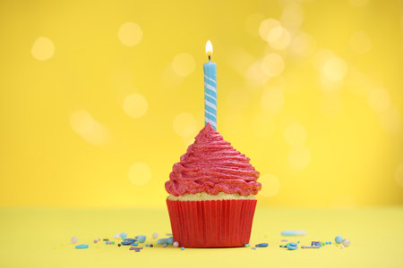 Burning candle in cupcake and sprinkles on yellow background with blurred lightsの写真素材