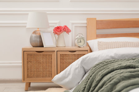 Bedside table with lamp, alarm clock and flowers near bed indoorsの写真素材