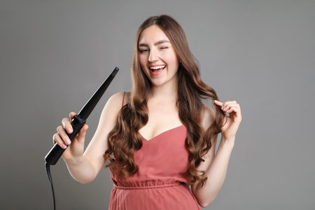 Woman with beautiful hairstyle and curling iron on grey backgroundの写真素材