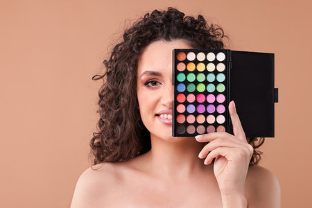 Smiling woman covering eye with eyeshadow palette on beige backgroundの写真素材