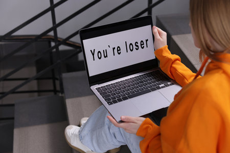 Cyber bullying. Upset teenage girl looking at laptop with text You Loser on stairs indoors, closeupの写真素材