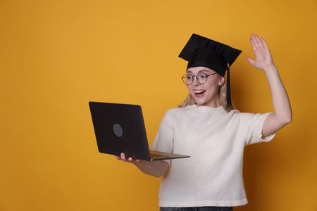 Happy student with laptop waving hello after graduation on orange backgroundの写真素材