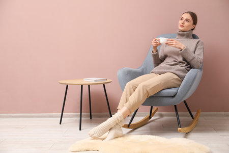 Beautiful woman with cup of drink relaxing in comfortable rocking chair near beige wall indoorsの写真素材