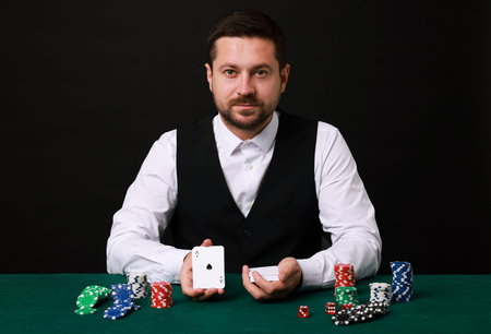 Professional croupier with playing cards at gambling table against black background