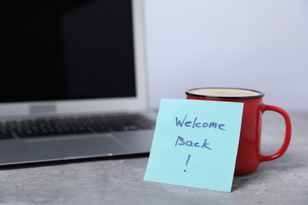 Sticky note with phrase Welcome Back, coffee and laptop on grey table, closeupの写真素材