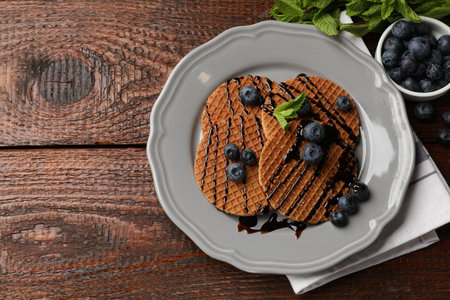 Tasty Dutch waffles (stroopwafels) with blueberries, mint and chocolate sauce on wooden table, flat lay. Space for textの写真素材