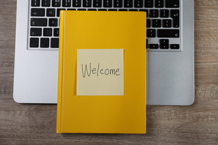 Sticky note with word Welcome, notebook and laptop on wooden table, top viewの写真素材