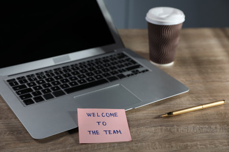 Paper note with words Welcome To The Team and laptop on wooden table, closeupの写真素材