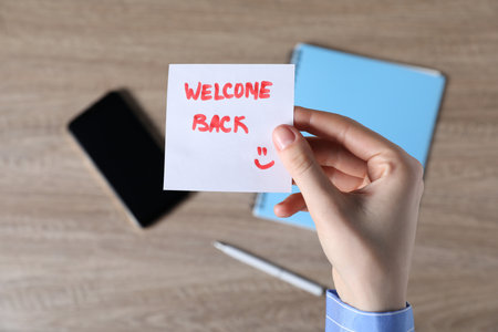 Woman holding paper note with words Welcome Back at wooden table, top viewの写真素材