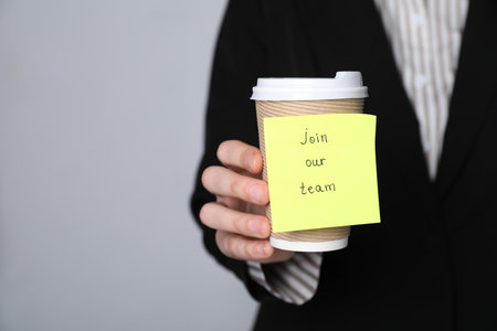 Join Our Team. Human resources manager holding takeaway cup with sticky note on grey background, closeup. Space for textの写真素材