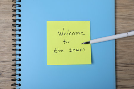 Sticky note with phrase Welcome To The Team, pen and notebook on wooden table, top viewの写真素材