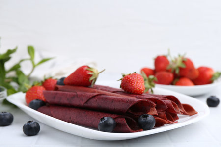 Tasty fruit leather rolls, strawberries and blueberries on white table, closeup. Space for text