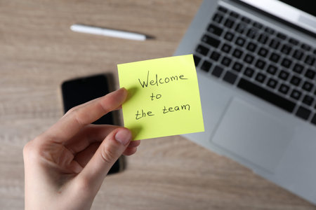 Woman holding paper note with words Welcome To The Team at wooden table, top view