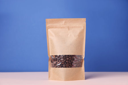 One zipper stand-up pouch with coffee beans on white table against blue background. Mockup for...