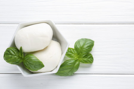 Pieces of delicious mozzarella cheese and basil in bowl on white wooden table, flat lay. Space for...