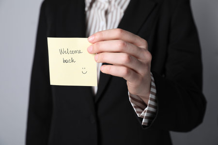 Businesswoman holding sticky note with word Welcome Back on gray background, closeup