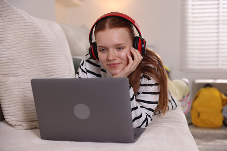 Teenage student in headphones using laptop while studying on sofa at homeの写真素材