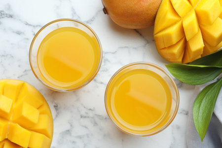Tasty mango juice in glasses, fresh fruits and green leaves on white marble table, flat lay