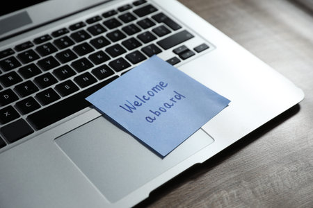 Paper note with words Welcome Aboard and laptop on wooden table, closeupの写真素材