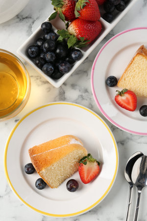 Pieces of tasty bundt cake with berries and tea on white marble table, flat lay
