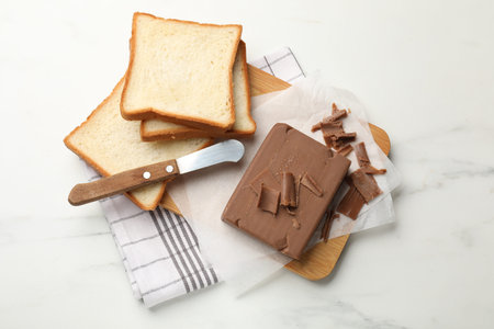 Block of tasty chocolate butter, bread slices and knife on white marble table, top viewの写真素材
