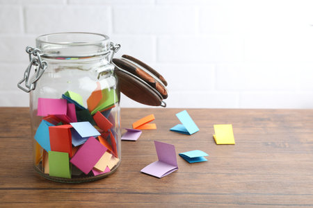 Jar with colorful pieces of paper on wooden table near white brick wall. Space for textの写真素材
