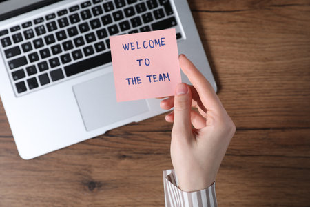 Woman holding paper note with words Welcome To The Team near laptop at wooden table, top viewの写真素材