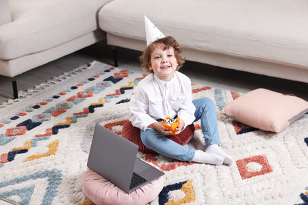 Birthday celebration. Little boy with cupcake having video call via laptop at home
