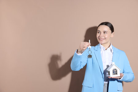 Real estate agent holding key with keychain and house model on beige background, space for textの写真素材