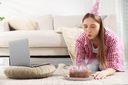 Birthday celebration. Woman blowing candles on cake while having video call via laptop at homeの写真素材