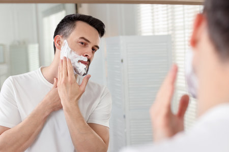 Handsome man applying shaving foam near mirror in bathroomの写真素材