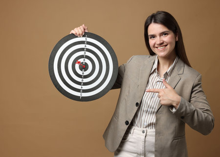 Targeting. Smiling businesswoman pointing at dart board and arrow on beige background