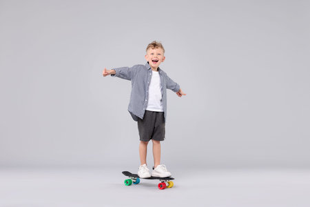 Cheerful little boy standing on penny board and showing thumbs up against gray background