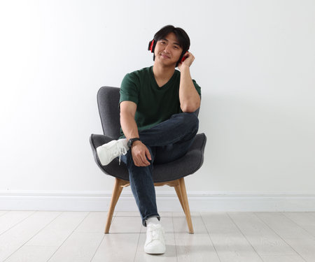 Handsome Asian man listening to music in headphones on armchair near white wallの写真素材