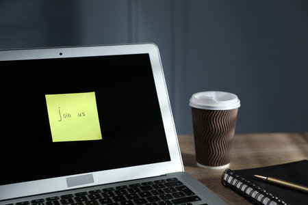 Sticky note with words Join Us, laptop, notepad and takeaway cup on wooden table against grey background, closeupの写真素材
