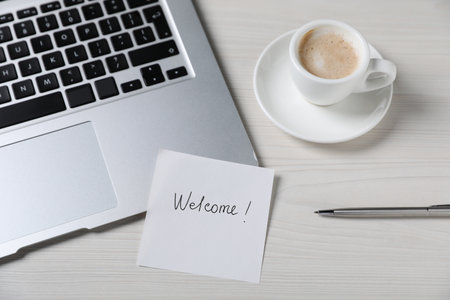 Paper note with word Welcome, laptop, cup of coffee and pen on light wooden table, above viewの写真素材