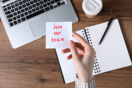 Woman holding paper note with words Join Our Team at wooden table, top view