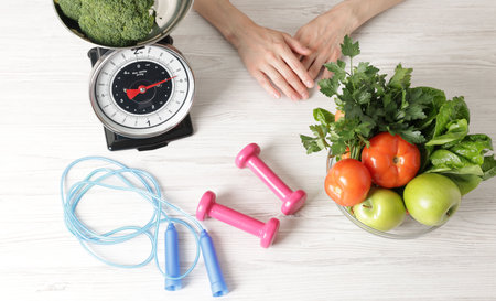 Woman at light wooden table with healthy food, kitchen scale, skipping rope and dumbbells, top viewの写真素材