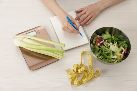 Woman developing diet plan at white wooden table with kitchen scale, healthy food and measuring tape, top viewの写真素材